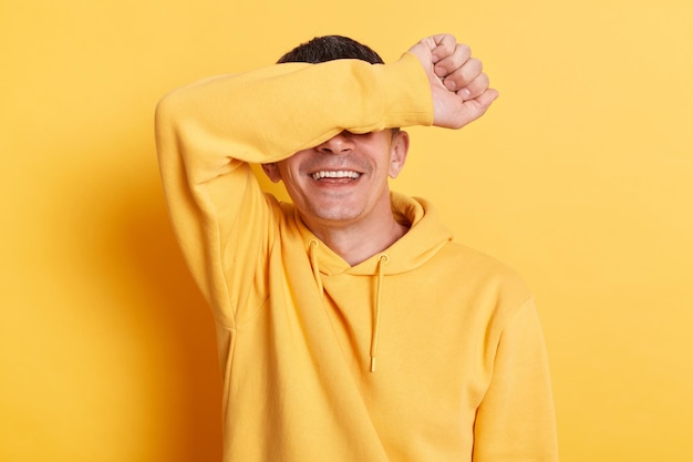 Binnenopname van lachende optimistische brunette man met hoodie die zijn ogen bedekt met arm, voelt verlegen en uiting gevend aan positieve emoties poseren geïsoleerd over gele achtergrond