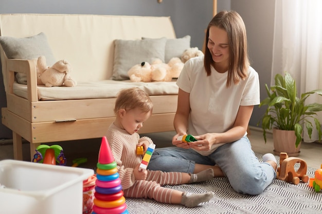 Binnenopname van moeder en babydochter die spelen met kleurrijke constructorblokken terwijl ze op de vloer zitten in de woonkamer thuis, familie die samen tijd doorbrengt