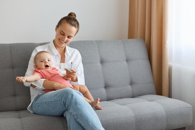 Binnenopname van mooie blanke vrouw met knotkapsel in wit overhemd en spijkerbroek zittend op de bank met baby en spelende mama die tijd doorbrengt met haar peuterkind