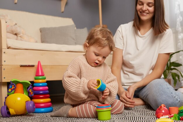 Binnenopname van mooie moeder speelt met baby in de kamer met kleurrijk plastic speelgoed gelukkige tijd samen vroege ontwikkeling mama geniet van haar zwangerschapsverlof