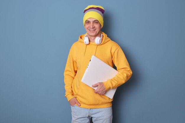 Binnenopname van opgetogen aantrekkelijke man met muts en casual gele hoodie die tegen de blauwe muur poseert en erg blij is te wachten op het begin van het werk en positieve emoties uitdrukt