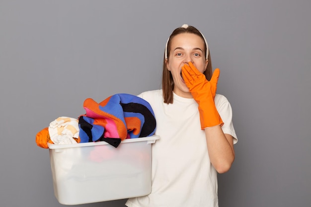 Binnenopname van opgewonden geschokte vrouw in wit t-shirt en haarband die een wasbak vasthoudt met kleding die huishoudelijke klusjes doet die de mond bedekken met palm die geïsoleerd staat over grijze achtergrond