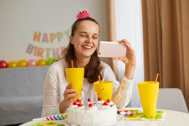 Binnenopname van optimistische positieve vrouw in witte kleding die aan tafel zit met cake en drankje, smartphone en glas vasthoudt met drankje, videogesprek heeft, online feest.