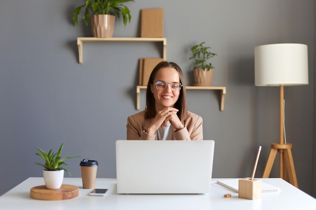 Binnenopname van schattige innemende vrouw vrouw met donker haar in beige jas en bril zittend aan tafel in kantoor werkend op laptop kijkend naar camera houdt handen onder de kin