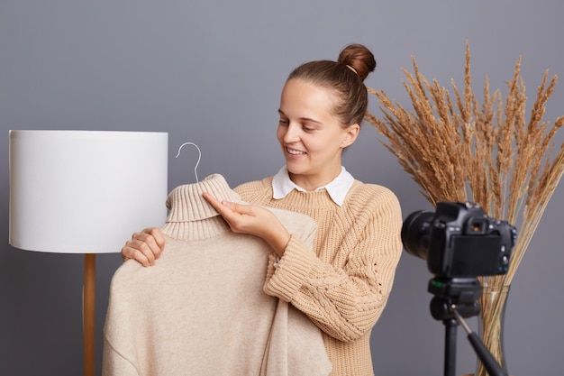 Binnenopname van tevreden gelukkige vrouw met beige trui tegen grijze muur met lamp en gedroogde bloemen die voor statief staan met camera-opnamevideo die haar kleding verkoopt