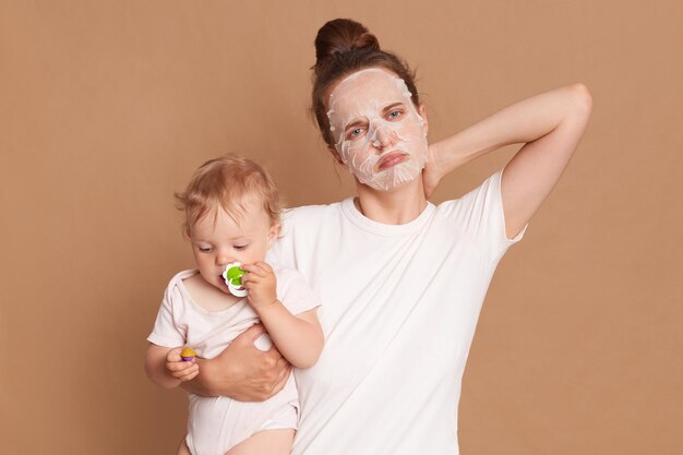 Binnenopname van trieste, ongezonde vrouw met een wit t-shirt die cosmetische ingrepen doet, voelt zich onwel heeft pijn in de nek die naar de camera kijkt met een droevige uitdrukking die babydochter in handen houdt