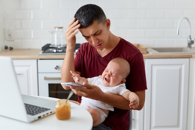 Binnenopname van trieste vader met een kastanjebruin casual t-shirt zittend met babyjongen of -meisje in de keuken, smartphone vasthoudend, het nummer van de vrouw bellend, kan dochter of zoon niet kalmeren.