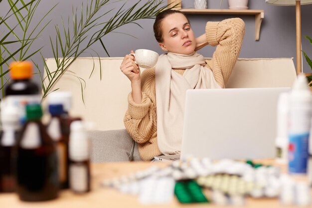 Binnenopname van vermoeide, uitgeputte, verveelde vrouw zittend op de bank gewikkeld in een sjaal omringd met medicijnen die thee drinkt terwijl ze haar nek aanraakt kijkend naar laptopscherm poserend in het leven