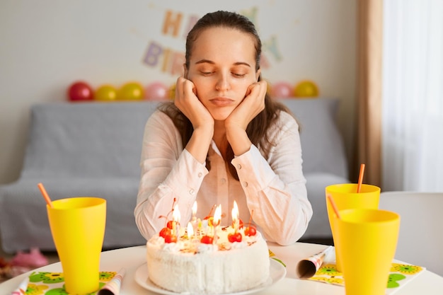 Binnenopname van verveelde, trieste blanke jonge volwassen vrouw die aan tafel zit en kijkt naar verjaardagstaart die boos is om alleen te vieren terwijl ze op haar gasten wacht