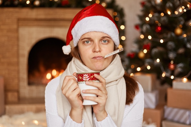 Binnenopname van zieke zieke vrouw met kerstmuts, gewikkeld in een warme sjaal, poserend met een thermometer in de mond, kijkend naar de camera, zittend in de buurt van de kerstboom en de open haard.