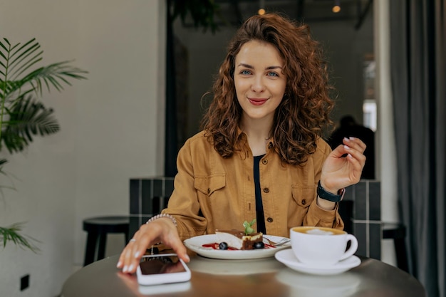 Binnenportret van een europees mooi glimlachend meisje met krullen met een bruin shirt dat 's ochtends ontbijt in een modern, stijlvol café