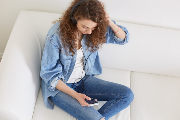 Binnenschot van aantrekkelijke dame met hoofdtelefoons, luisterend aan muziek, hoogste mening van geconcentreerd ontspannen model, zittend op bank, genietend van haar vrije tijd, dragend vrijetijdskleding. mensen en rust concept.