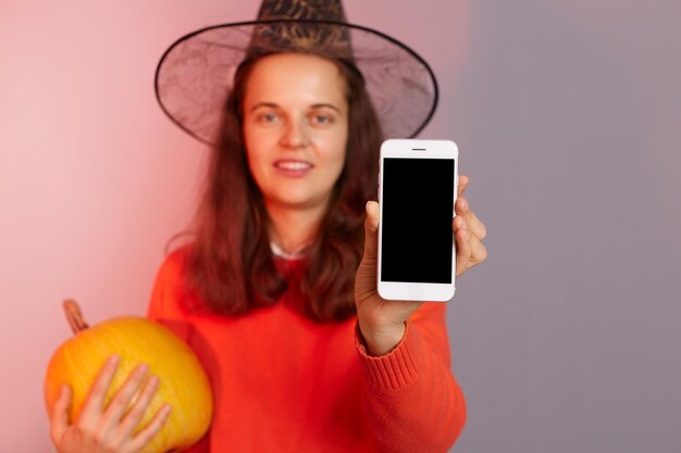 Binnenschot van aantrekkelijke vrouw die heksenhoed draagt die zich met oranje pompoen in handen bevindt en mobiele telefoon toont met lege vertoning die zich geïsoleerd over neonlichtachtergrond viert die Halloween viert