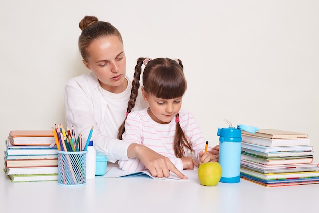 Binnenschot van geconcentreerde vrouwelijke leraar die met haar leerling aan het bureau zit en taken op school doet, zittend aan het bureau tussen boeken geïsoleerd op witte achtergrond