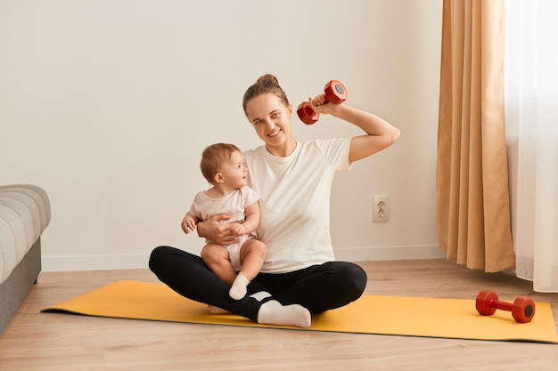 Binnenschot van glimlachende vrouw die wit t-shirt en zwarte leggins draagt die sportoefeningen doen met baby in handen die op yogamat zitten en hand opsteken met halter in handen die biceps trainen