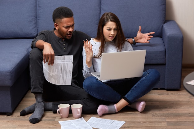 Binnenschot van multicultureel paar die vrije tijd thuis doorbrengen en terloops gekleed rekeningen controleren, zittend voor laptop