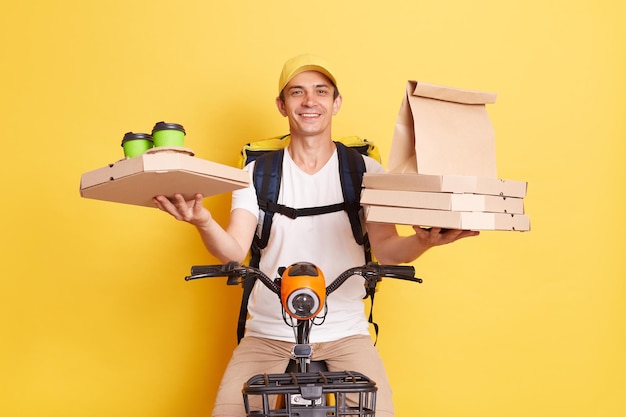 Binnenschot van positieve optimistische koerier met pet en wit t-shirt met pizzadozen en koffie van koffie die op de fiets zit en voedsel levert geïsoleerd op gele achtergrond