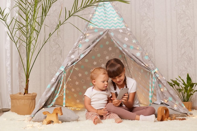 Binnenschot van schattige positieve gelukkige broers en zussen die witte t-shirts dragen die in een peetee-tent zitten en een mobiele telefoon gebruiken die met een glimlach naar de weergave van de smartphone kijkt