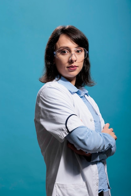 Biochemistry specialist wearing protective goggles and lab coat standing on blue background while looking at camera. Biomedical researcher with crossed arms standing on blue background, Studio shot.