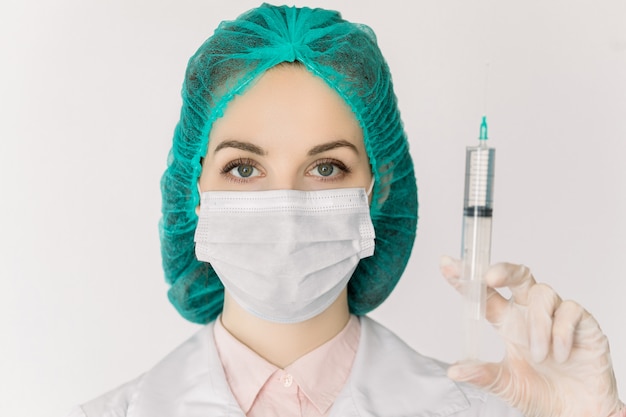 Biological hazard, epidemic of coronavirus, covid-19, flu, TB. Young Caucasian doctor woman in protective cap and mask holds an injection syringe with vaccine or medicine, isolated on white background