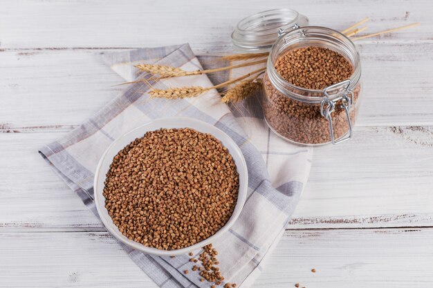 Biologische ongekookte verspreide boekweitkorrel in een kom en glazen pot op een witte houten rustiek. Gezond en dieetvoedselconcept.