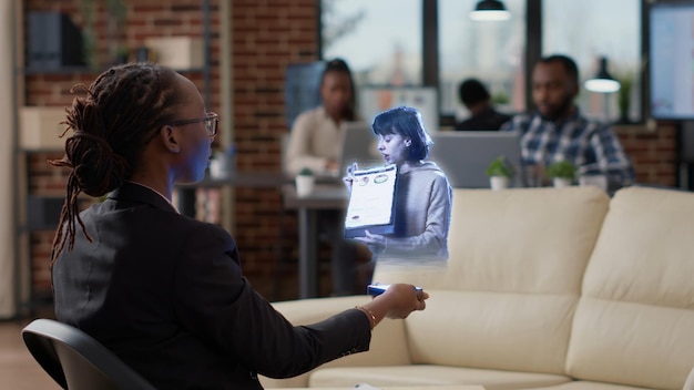 BIPOC employee in augmented reality teleconference meeting in high tech office talking with coworker. Accountant at workplace desk in online videocall with manager using hologram technology