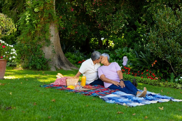 Biraciaal senior paar zoenen zittend met eten en drinken op deken tegen planten in het park