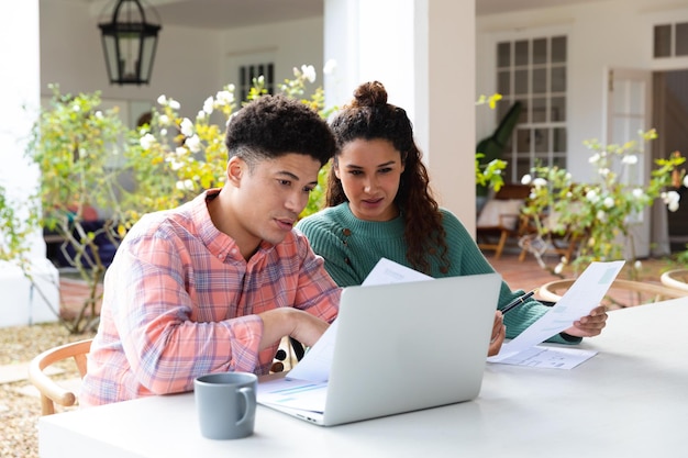 Biracial couple sitting on garden terrace outside house using laptop and paying bills. Inclusivity, domestic life and togetherness concept.