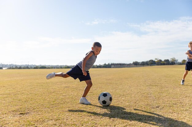 Biracial elementary schoolboy kicking soccer ball while playing on field against sky