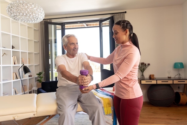 Photo biracial female physiotherapist helping caucasian senior man lifting dumbbell while sitting at home