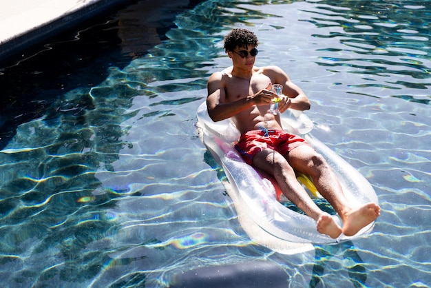 선글라스를 쓴 혼혈인 남성은 수영장에서 칵테일을 마시며 풍선 위에서 휴식을 취하며 공간을 복사합니다. 여름, 자유 시간, 휴식 및 휴가.