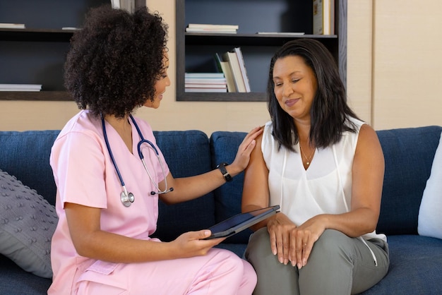biracial nurse holding tablet is talking to mature biracial woman at home