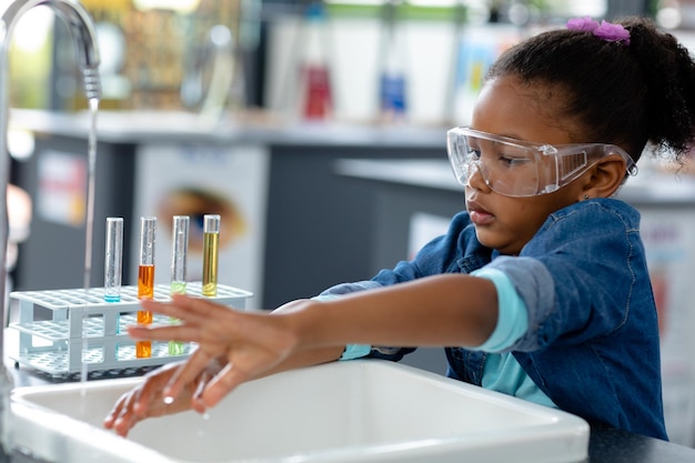 Biracial schoolgirl in safety glasses washing hands in science class