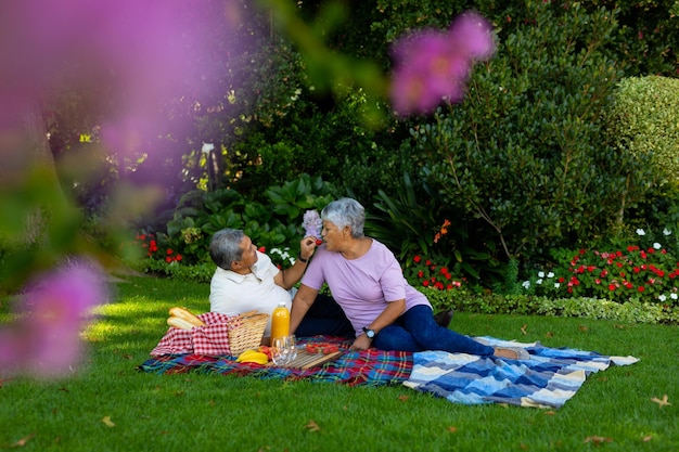 Biracial senior man feeding strawberry to wife while sitting on blanket against plants in park