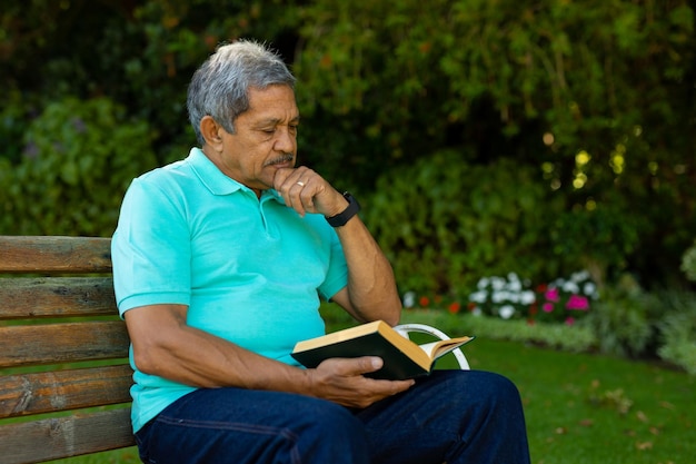 Biracial senior man with hand on chin reading book while sitting on bench against plants in park