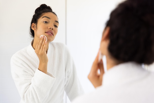 Photo biracial woman in bathrobe cleansing her face in sunny bathroom