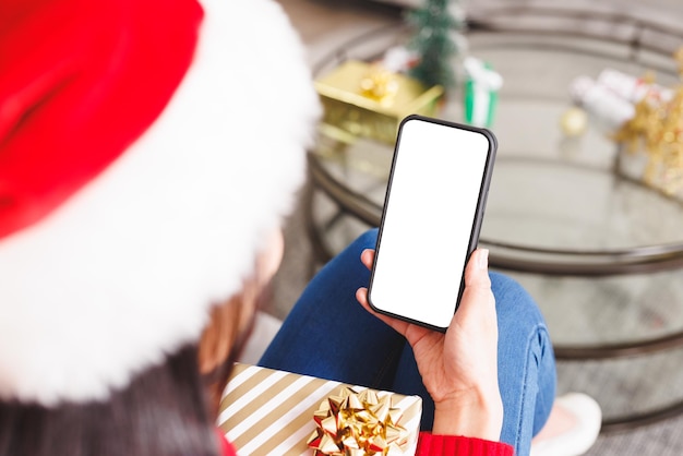 Biracial woman wearing santa hat, sitting on sofa in living room, using smartphone with copyspace