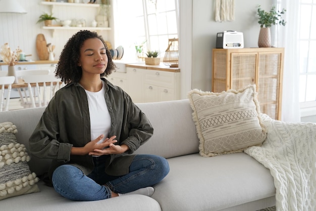 Biracial young girl enjoy meditation practice yoga sitting on sofa in lotus pose at home