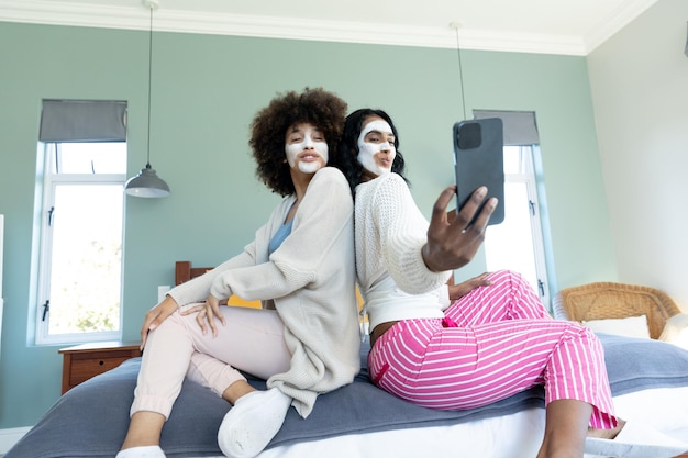 Biraciale vrienden met crème op gezichten poseren en selfie nemen via smartphone terwijl ze op bed zitten. Technologie, gelukkig, huidverzorging, verwennerij, onveranderd, vriendschap, samenzijn, levensstijl en thuis.