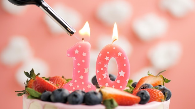 Birthday cake number 10 pink candle on beautiful cake with berries and lighter with fire against background of white clouds and pink sky Closeup view