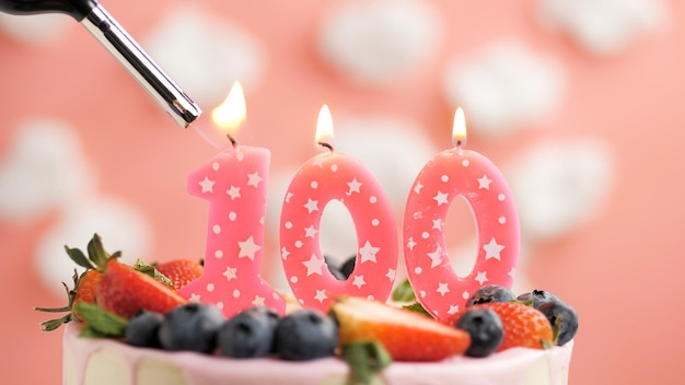 Birthday cake number 100 pink candle on beautiful cake with berries and lighter with fire against background of white clouds and pink sky Closeup view