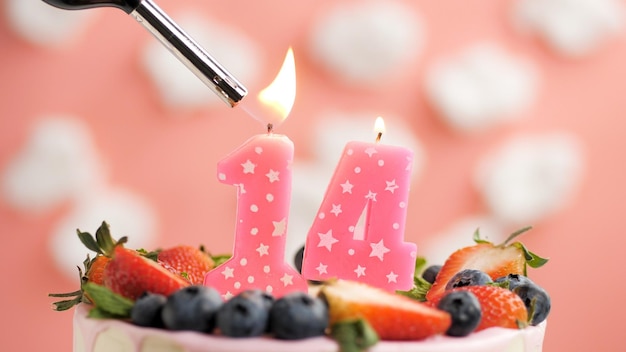 Birthday cake number 14 pink candle on beautiful cake with berries and lighter with fire against background of white clouds and pink sky Closeup view