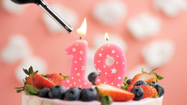 Birthday cake number 19 pink candle on beautiful cake with berries and lighter with fire against background of white clouds and pink sky Closeup view