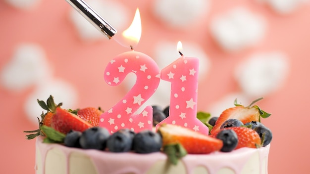 Birthday cake number 21 pink candle on beautiful cake with\
berries and lighter with fire against background of white clouds\
and pink sky closeup view