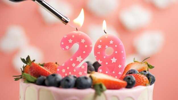 Birthday cake number 29 pink candle on beautiful cake with\
berries and lighter with fire against background of white clouds\
and pink sky closeup view