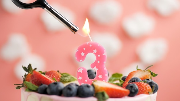 Birthday cake number 3 pink candle on beautiful cake with\
berries and lighter with fire against background of white clouds\
and pink sky closeup view