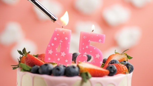 Birthday cake number 45 pink candle on beautiful cake with\
berries and lighter with fire against background of white clouds\
and pink sky closeup view