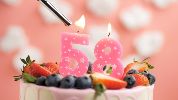 Birthday cake number 58 pink candle on beautiful cake with\
berries and lighter with fire against background of white clouds\
and pink sky closeup view