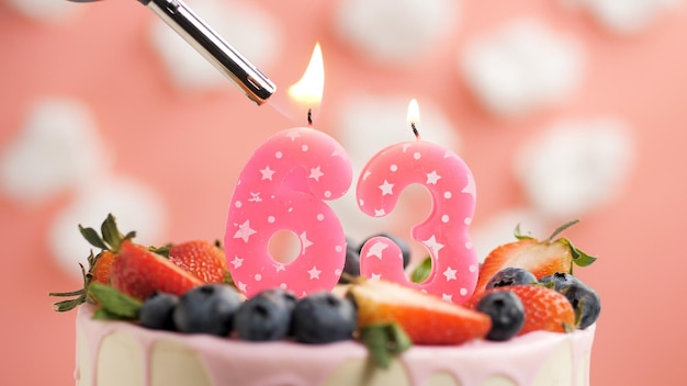 Birthday cake number 63 pink candle on beautiful cake with
berries and lighter with fire against background of white clouds
and pink sky closeup view