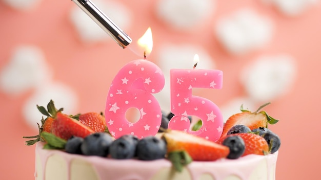 Birthday cake number 65 pink candle on beautiful cake with\
berries and lighter with fire against background of white clouds\
and pink sky closeup view